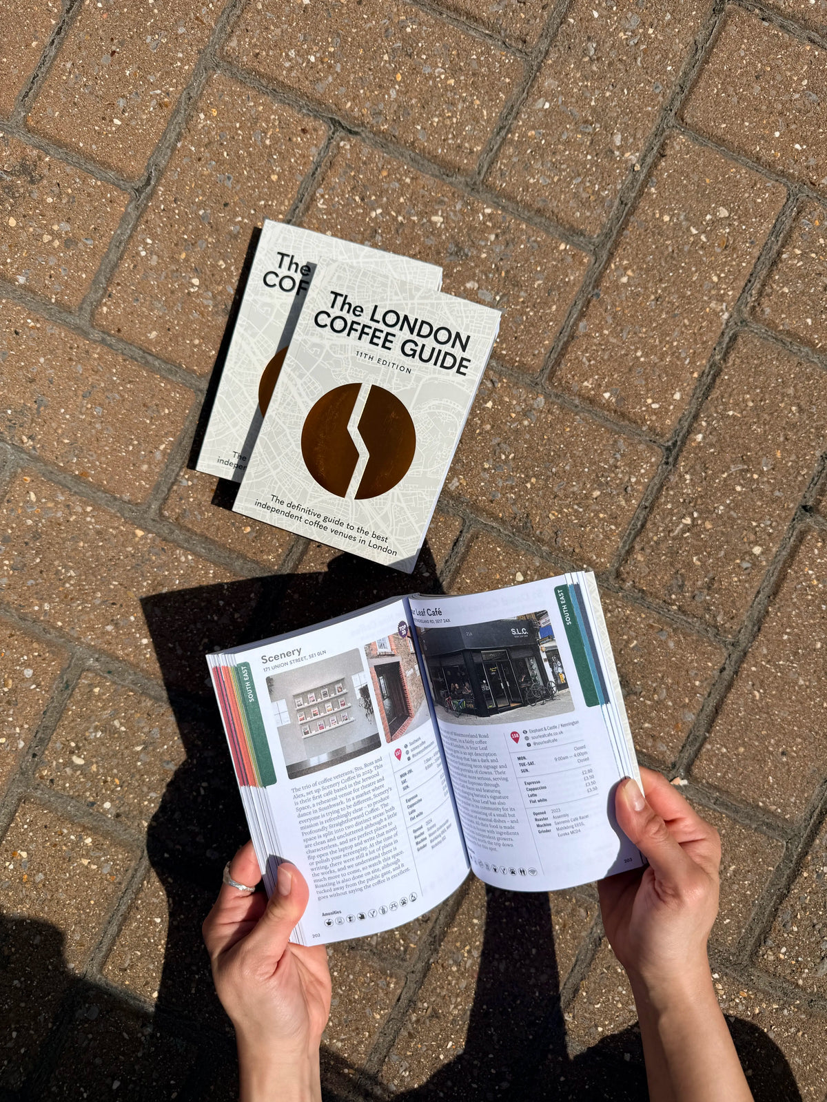 Person holding 'The London Coffee Guide' on a brick pavement