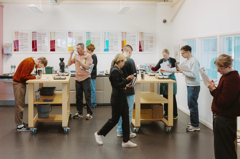 A group of people gathered around wooden worktables in a bright, modern indoor workshop or classroom with white walls, posters on the wall, and large windows on the right.