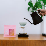 Person pouring coffee into a filter with a pink coffee bag labeled 'PLOT' on a wooden surface.