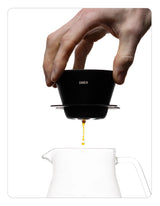 Hand pouring coffee from a OREA 01 Obsidian coated steel brewer into a clear glass pot on a white background